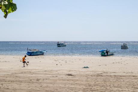 A boy kicks a ball on a beach with fishing boats